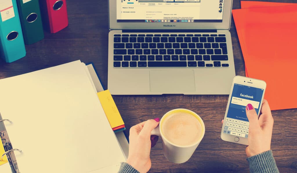 Social Media for care homes -Woman sitting at desk holding iphone with coffee and macbook
