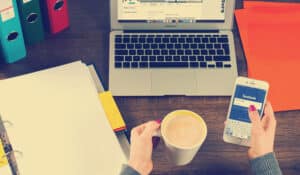 Social Media for care homes -Woman sitting at desk holding iphone with coffee and macbook