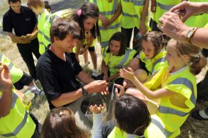 A man demonstrates an activity to a group of children, all wearing bright yellow safety vests, seated on grass in a sunny outdoor setting.