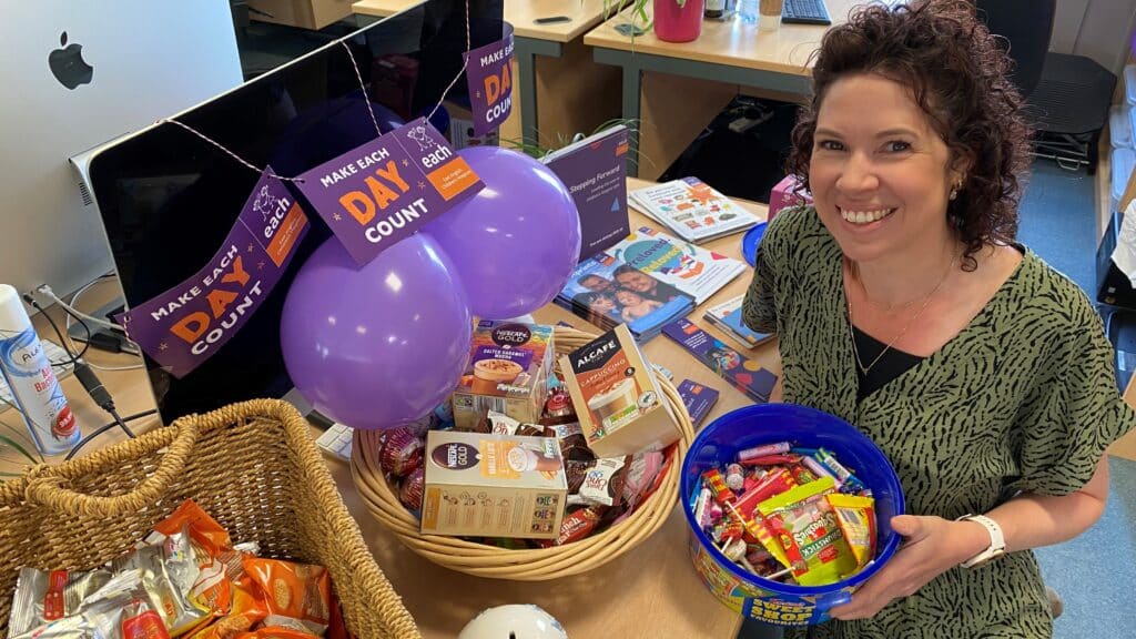 A woman with curly hair smiles while holding a blue bowl filled with candy, beside a basket of snacks and purple balloons, in a vibrant office setting.