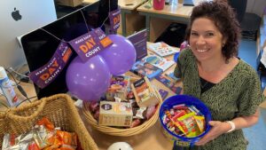 A woman with curly hair smiles while holding a blue bowl filled with candy, beside a basket of snacks and purple balloons, in a vibrant office setting.