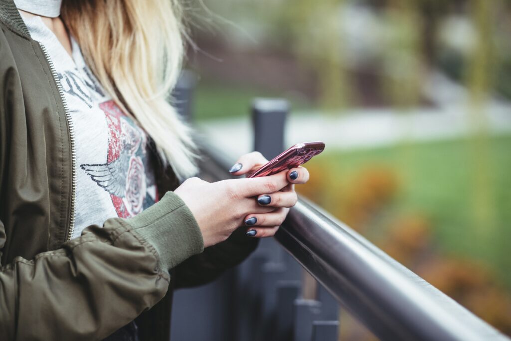 A person wearing a graphic t-shirt and olive jacket holds a pink smartphone while leaning on a black railing, with greenery in the background.