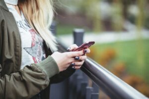 A person wearing a graphic t-shirt and olive jacket holds a pink smartphone while leaning on a black railing, with greenery in the background.