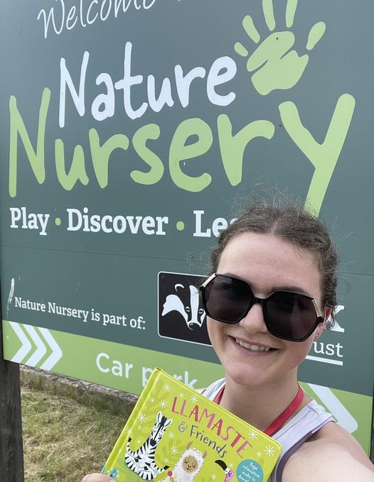 Smiling individual holding a children's book stands in front of a green sign that reads "Nature Nursery," highlighting play and discovery.