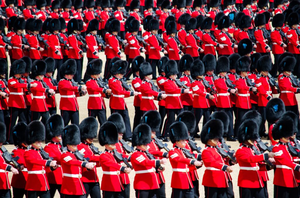 A parade of soldiers in bright red uniforms and tall black fur hats, standing in formation, holding rifles, against a neutral background.
