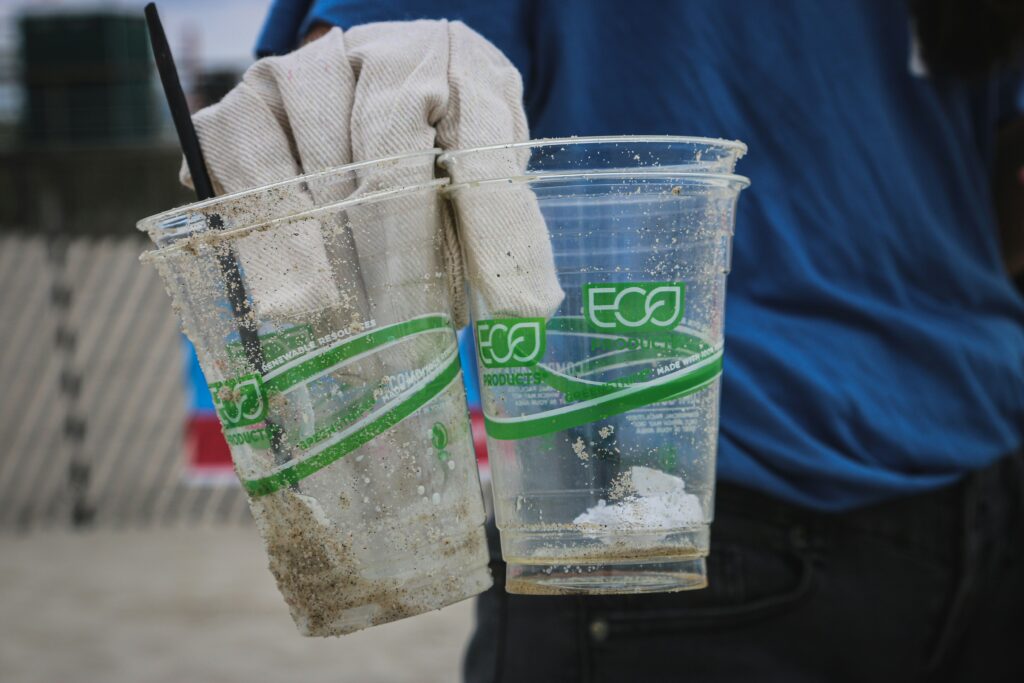 Two clear, eco-friendly cups filled with sand and debris are held by a gloved hand, with a person in blue behind them.