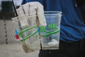 Two clear, eco-friendly cups filled with sand and debris are held by a gloved hand, with a person in blue behind them.