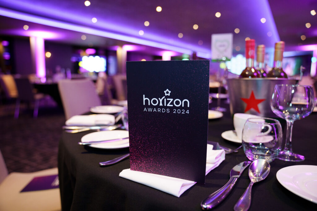 Horizon Awards 2024 menu stands on a elegantly set table with black tablecloth, cutlery, wine glasses, and a champagne bucket in a softly lit venue.