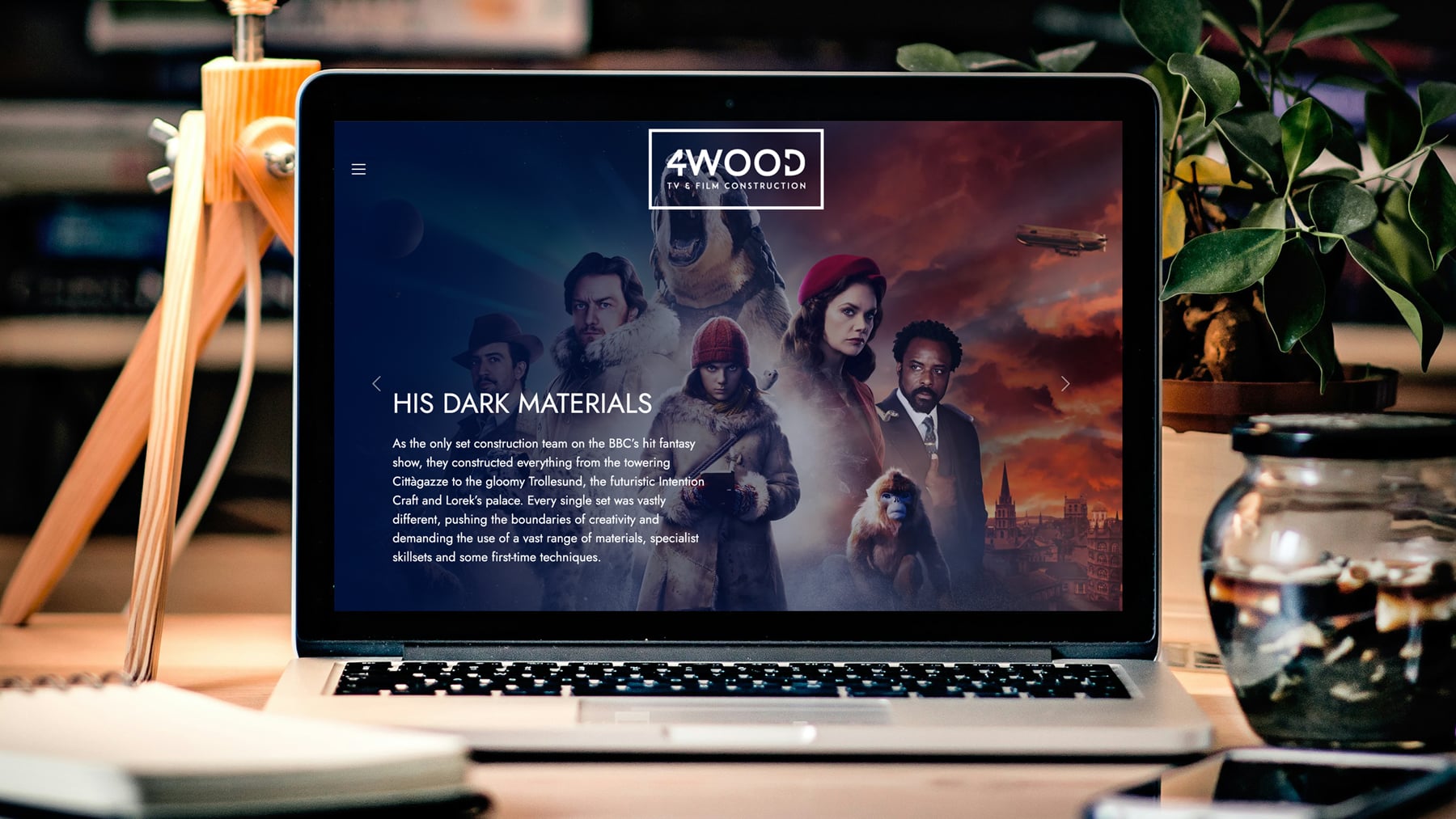 Laptop displaying "His Dark Materials" with a colorful background featuring characters from the series. A desk setting with a plant and a jar is visible.