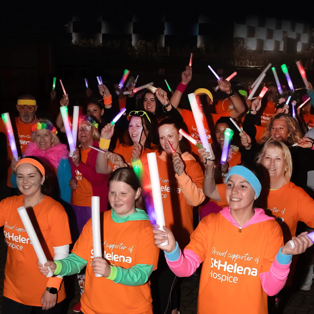 A crowd of supporters wearing bright orange shirts cheerfully wave colorful glow sticks, celebrating St. Helena Hospice under night lighting.