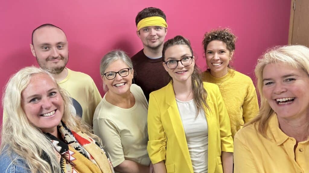 Group of seven smiling individuals in yellow outfits against a vibrant pink background, promoting mental health awareness.