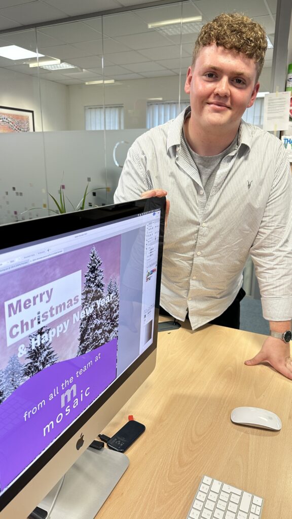 Man in a striped shirt smiles beside a computer displaying a festive graphic with "Merry Christmas & Happy New Year" and trees.
