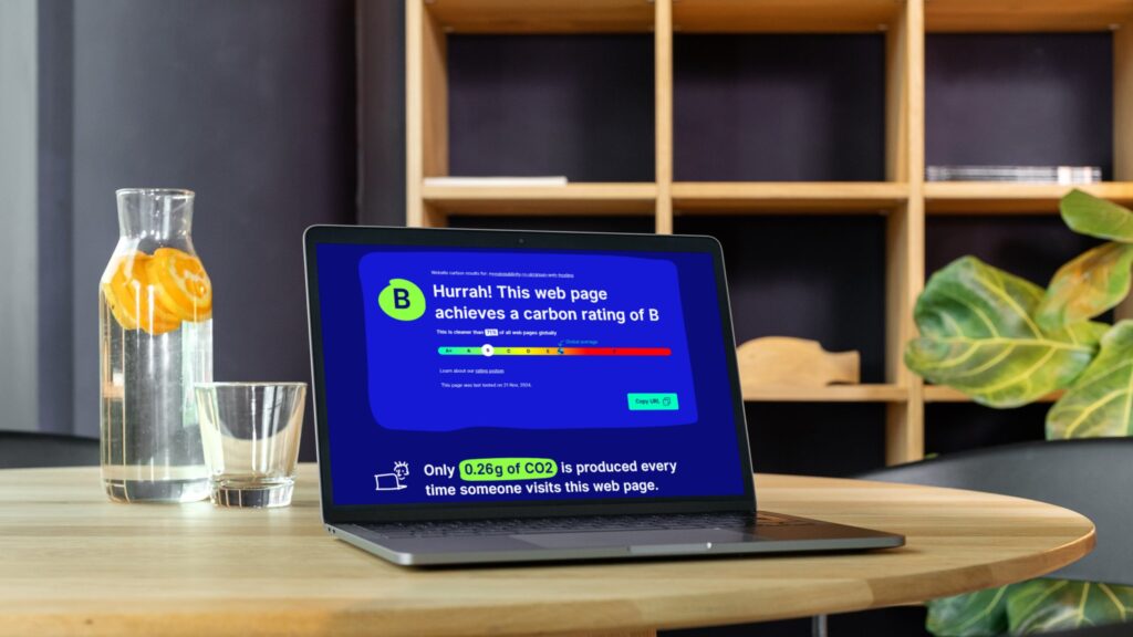 Laptop on a wooden table displays a vibrant blue screen with a carbon rating of B, beside a glass of water and a leafy plant.