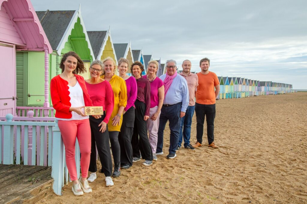 A group of nine people stands on a sandy beach, smiling, in front of colorful beach huts. The sky is overcast, reflecting a coastal atmosphere.