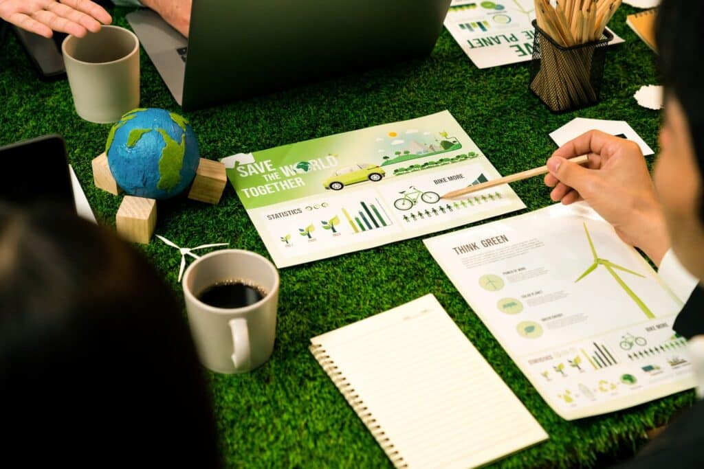 Hands and notebooks rest on a green, grass-like surface, featuring a globe, coffee cup, and illustrated documents promoting eco-friendly initiatives.