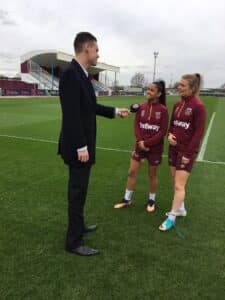 A man in a suit interviews two women in maroon sports jerseys on a grass pitch, with a cloudy sky and stadium in the background.