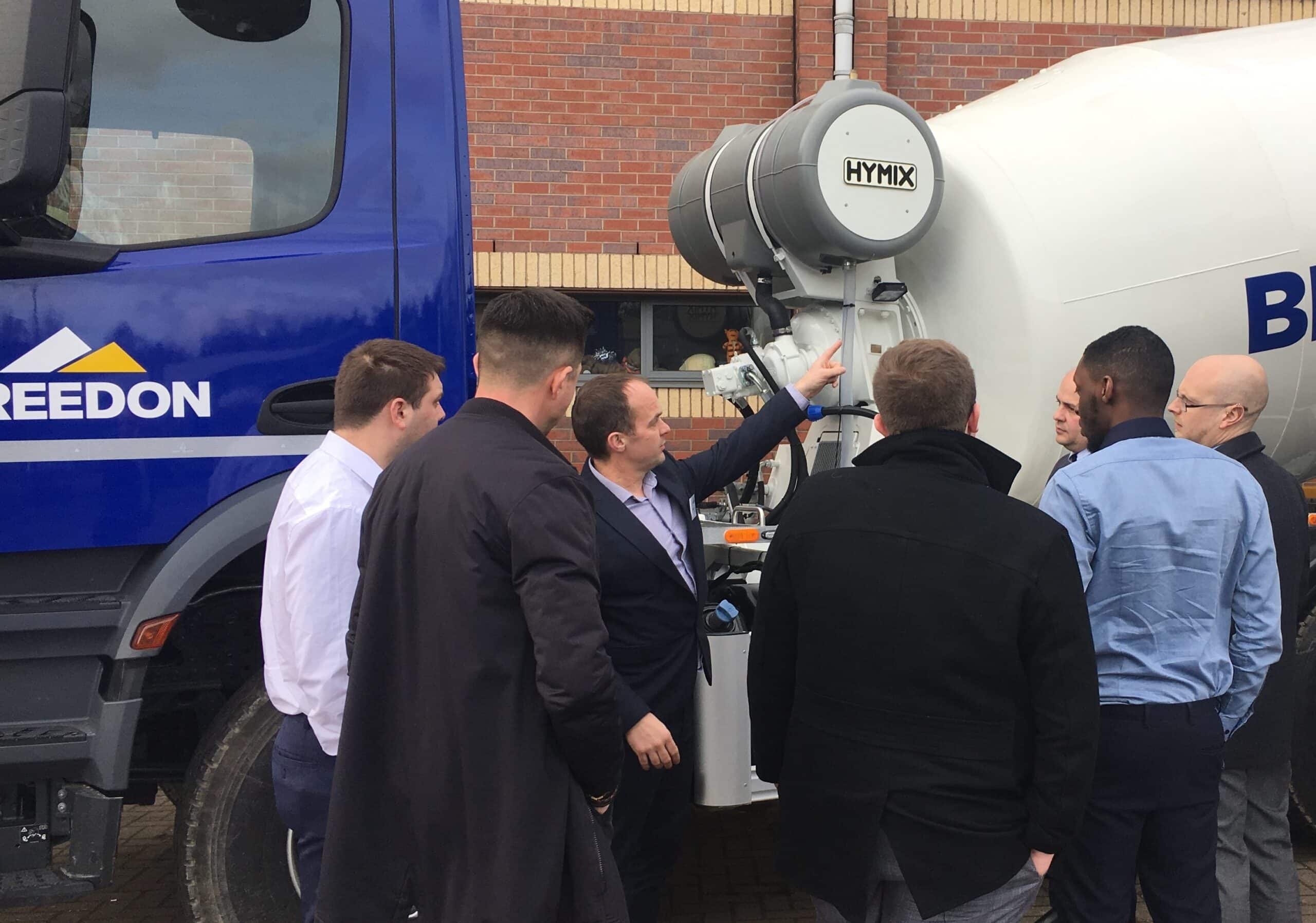 A group of six men gathers around a concrete mixer truck, discussing its features, with a brick building in the background.