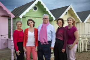 Five individuals stand in front of colorful beach huts, wearing shades of pink and red, showcasing a vibrant and cheerful atmosphere.