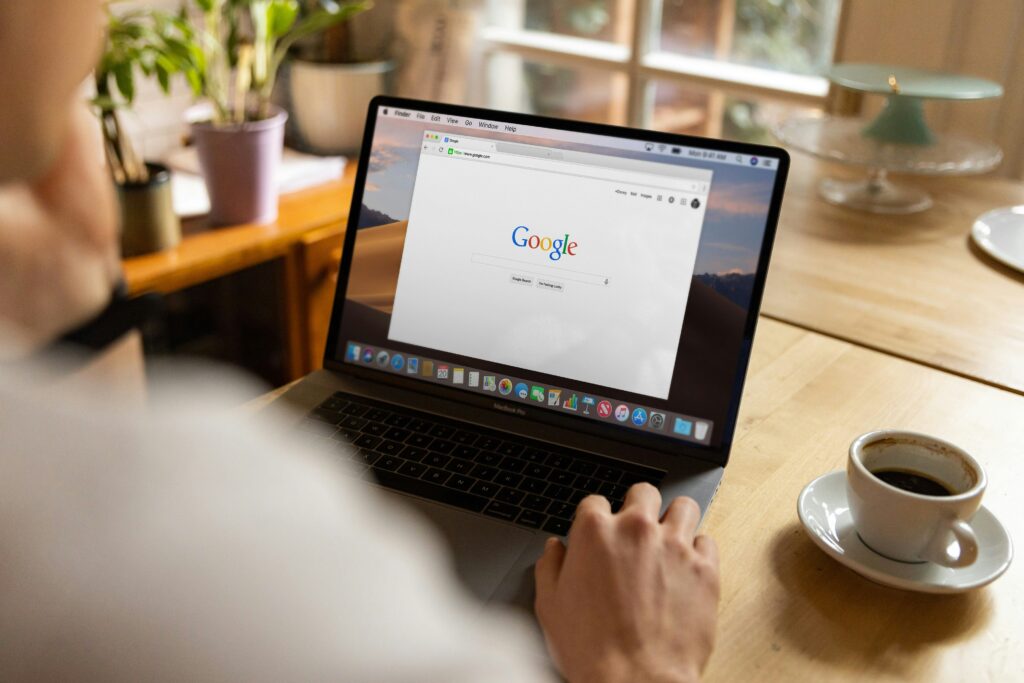 Person using a laptop displaying the Google homepage, with a cup of coffee on a wooden table and houseplants in the background.
