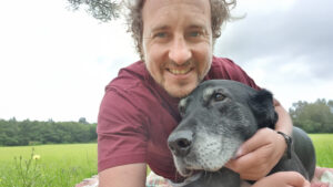 Smiling man in a maroon shirt embraces a black dog, both posing on a blanket in a lush green field under cloudy skies.