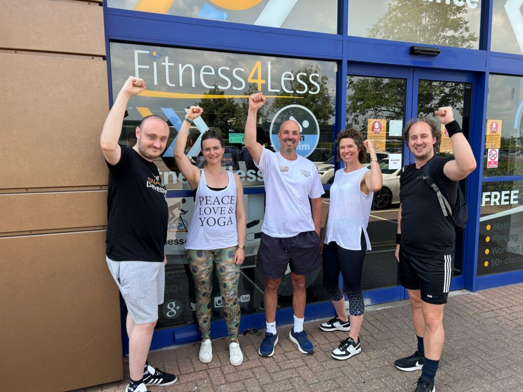 Five smiling individuals stand in front of the Fitness4Less gym, raising their fists in celebration. They wear athletic clothing in a variety of colors and patterns.