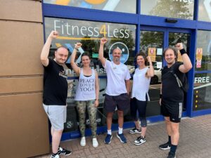 Five smiling individuals stand in front of the Fitness4Less gym, raising their fists in celebration. They wear athletic clothing in a variety of colors and patterns.