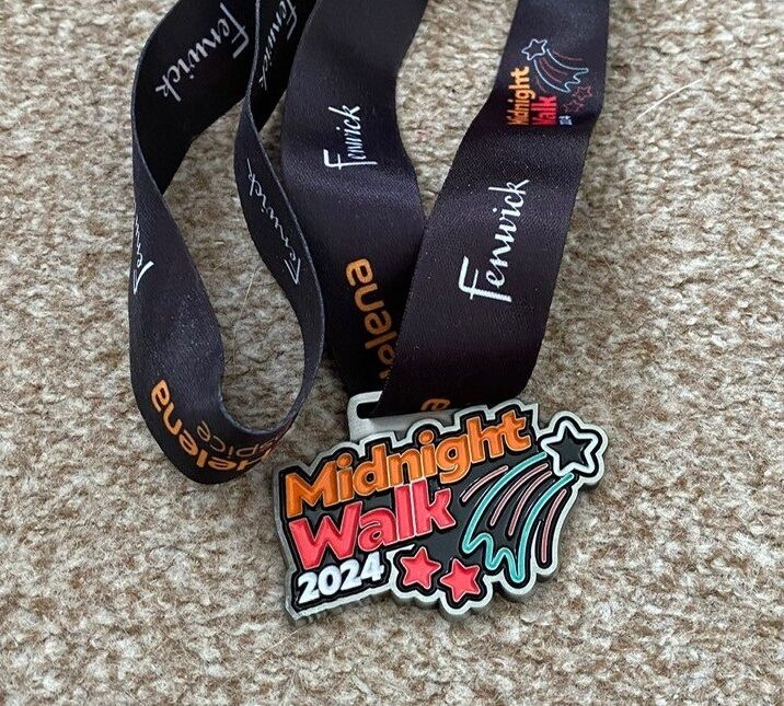A colorful medal with "Midnight Walk 2024" text, attached to a black ribbon featuring logos, resting on a textured beige surface.