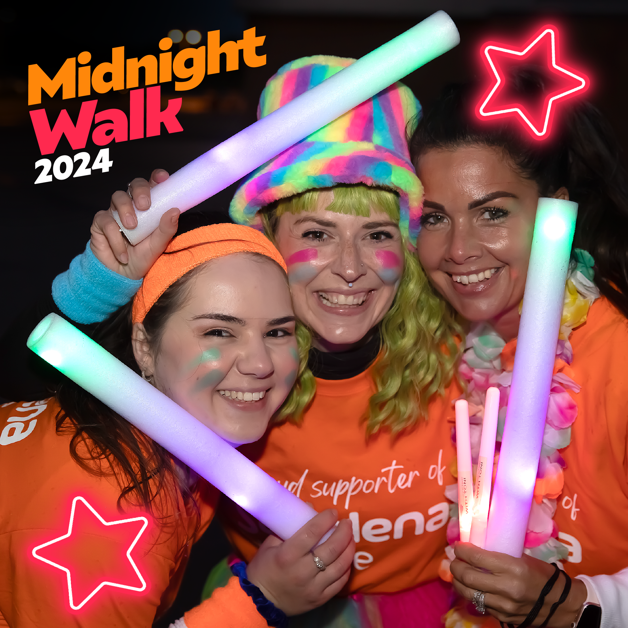 Three smiling participants in bright orange shirts hold glowing sticks, surrounded by colorful accessories and nighttime ambience.