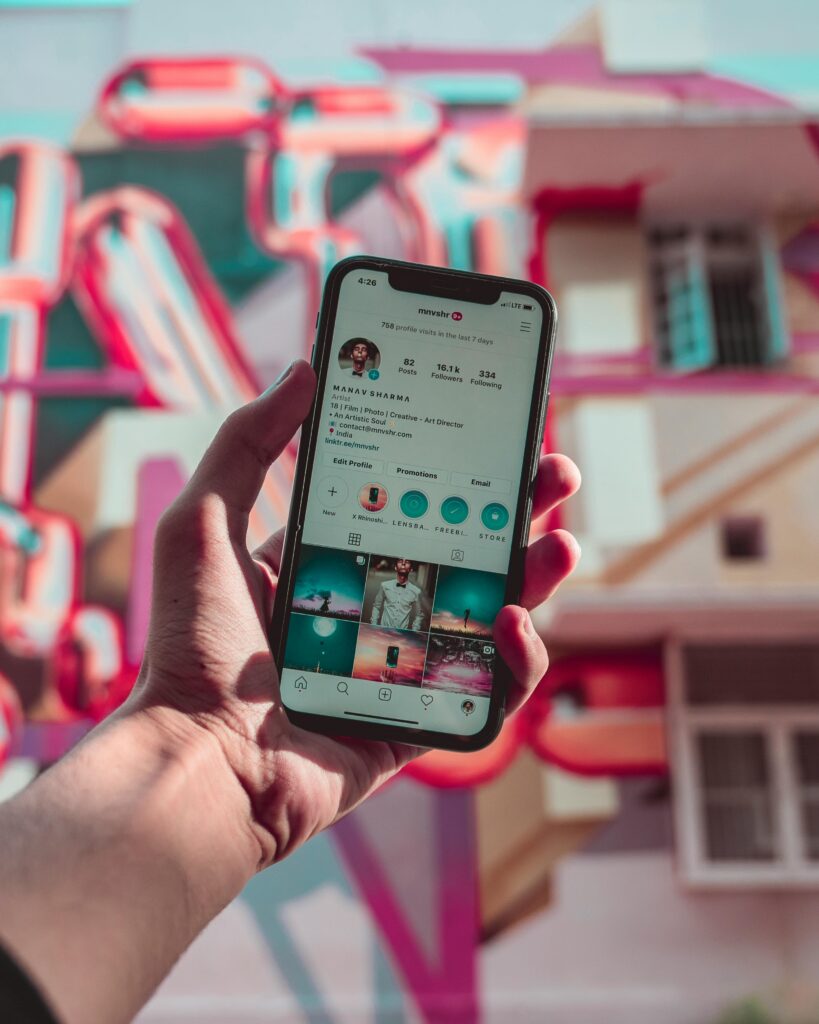 A hand holds a smartphone displaying an Instagram profile in front of a colorful mural with pink and blue elements.