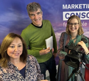 Three professionals pose in front of a purple backdrop, smiling. One holds a camera, another a notepad, and they wear vibrant clothing.