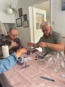 Two men are engaged in a creative activity at a table covered with a pink patterned cloth, surrounded by paint supplies and empty glasses.