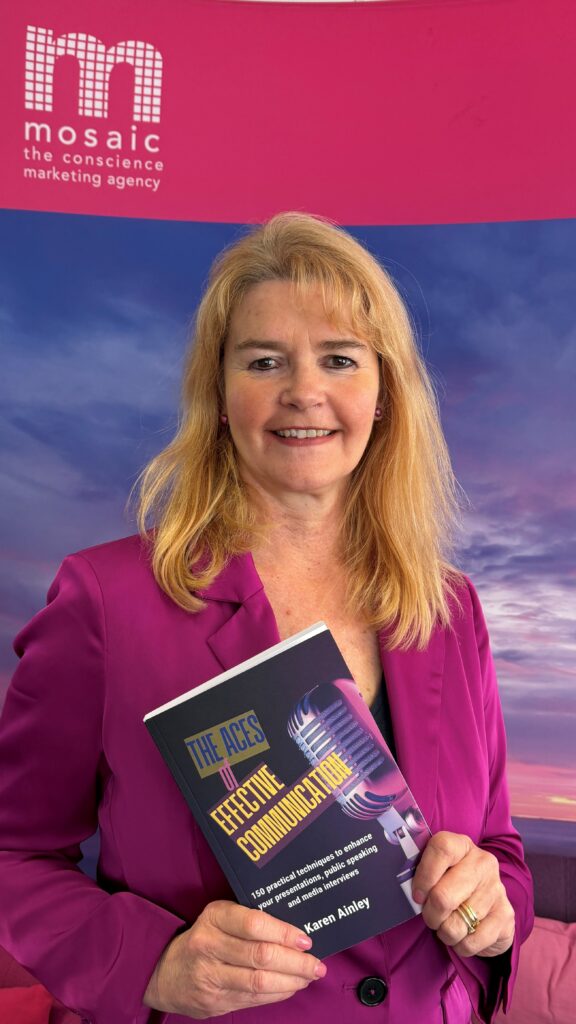 A woman with long blonde hair in a bright pink blazer holds a book titled "The Aces of Effective Communication" against a purple background.