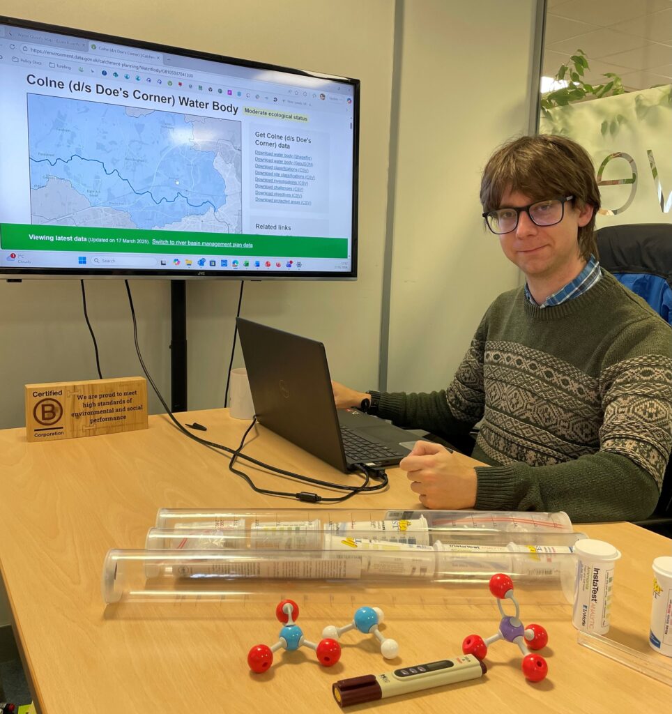 A person with brown hair and glasses sits at a desk with a laptop, displaying a water body map, surrounded by lab equipment and models.