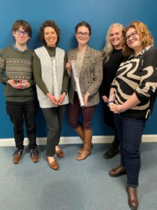 Five smiling individuals stand against a blue wall, holding various items. They wear smart-casual attire, with warm colours and textures.