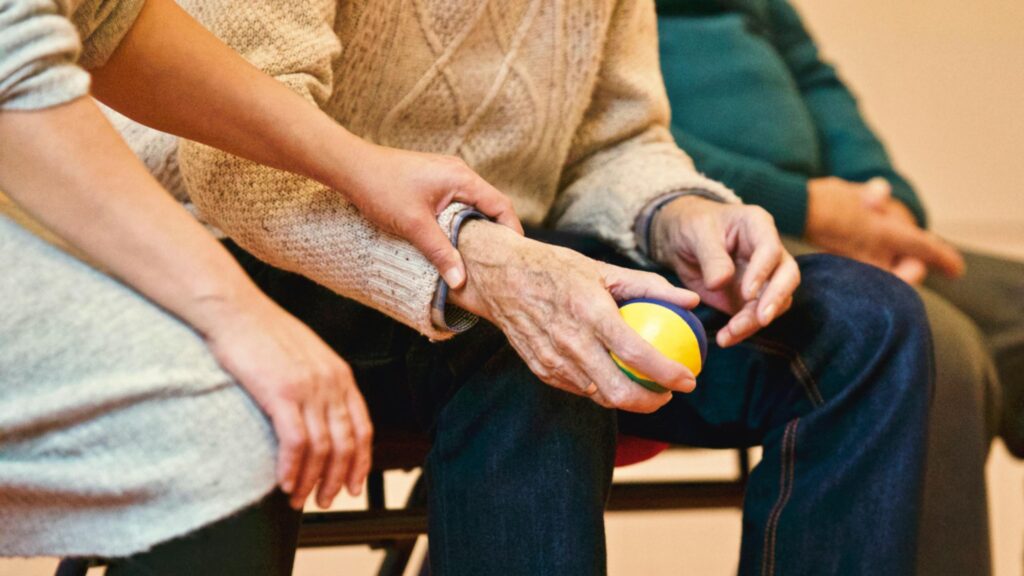 An elderly person holds a colourful stress ball, with a caregiver’s hand gently placed on their arm, both seated together.