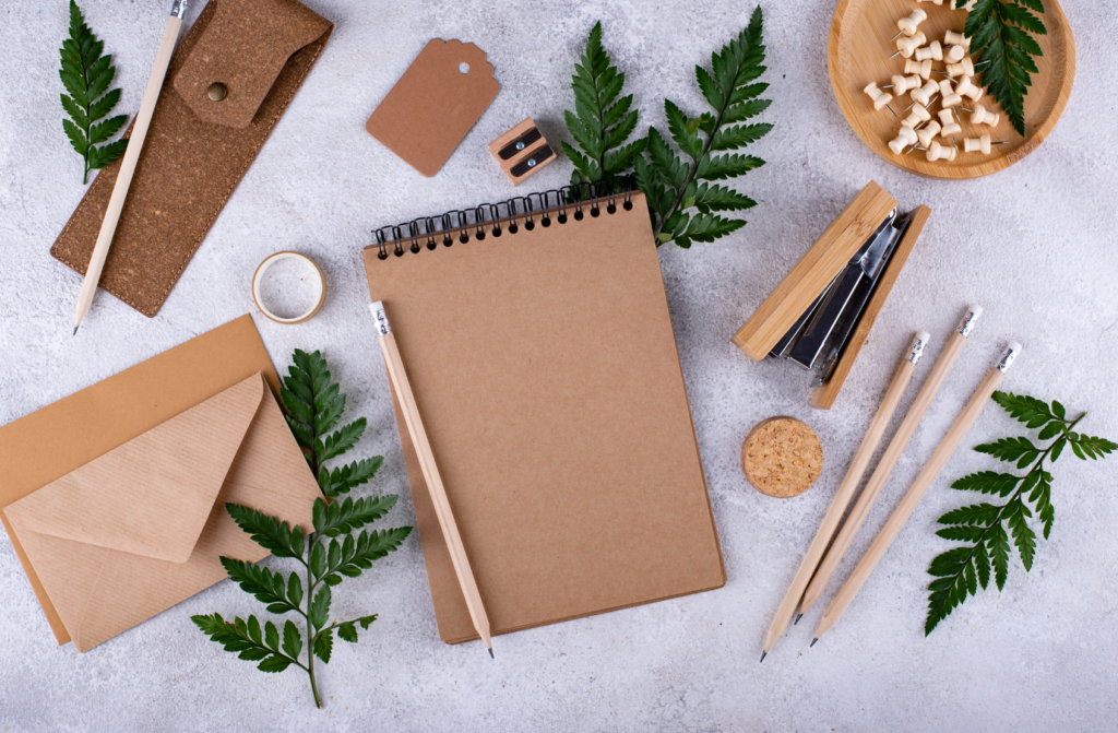 Flat lay of a brown notepad, pencils, an envelope, and natural elements like ferns and wooden pieces on a grey surface.