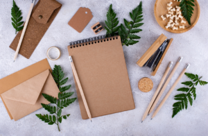 Flat lay of a brown notepad, pencils, an envelope, and natural elements like ferns and wooden pieces on a grey surface.