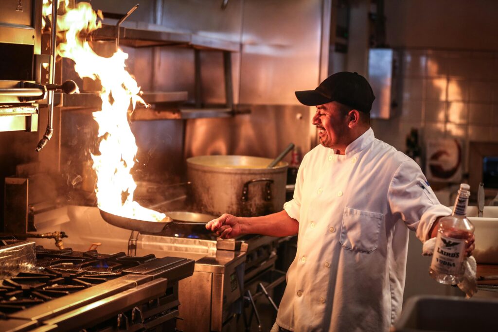Chef in a white coat, expertly tossing a flaming pan in a bustling kitchen, with sleek stainless steel appliances and pots in the background.
