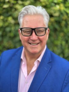 Smiling man with grey hair and glasses, wearing a blue blazer over a pink shirt, set against a green foliage background.